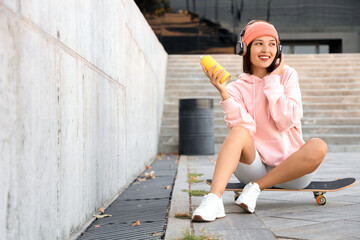 Young Asian woman in headphones with spray paint can sitting on skateboard outdoors