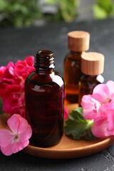 Bottles of geranium essential oil and beautiful flowers on black table, closeup