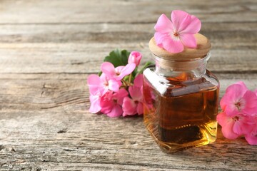 Bottle of geranium essential oil and beautiful flowers on wooden table, closeup. Space for text