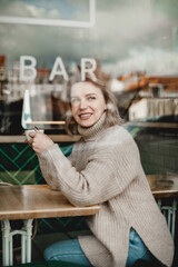 A woman is sitting at a table in a cafe, smiling and holding a cup of coffee. Concept of relaxation and enjoyment, as the woman takes a break from her day to savor her beverage