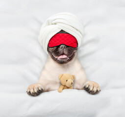 Pug puppy wearing sleeping mask sleeps with toy bear under white blanket on a bed at home. Top down...