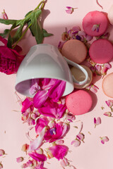 macaroni cookies on the table, next to a tea cup. Flower petals are falling out of the cup. Pink tablecloth and flowers on the table.