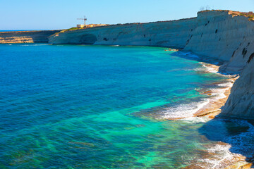 View of the coast of the island of Malta. Sea is calm and blue, with a rocky shoreline in the background. The water is clear and the waves are gentle