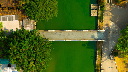 An aerial view captures the serene beauty of a footbridge over tranquil green waters, bordered by lush trees and urban structures