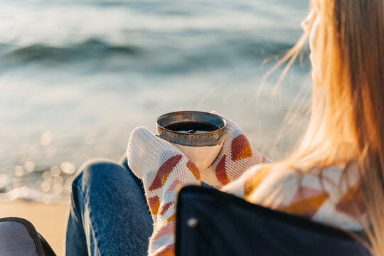 Closeup of a woman sitting in chair on winter sea shore in a cozy sweater and holding hot cup of coffee. A girl enjoying beautiful view of autumn ocean seaside waterfront waves.