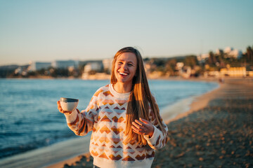 Young beautiful girl in cozy sweater holding coffee cup and smiling into the camera on seaside shore during mild sunset. Cute attractive woman enjoying her cup tea with autumn ocean waves background.