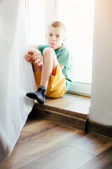 Cheerful school-age boy in a green T-shirt sitting at the window at home. Front view