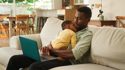 A Caring Father Working on Laptop While Holding His Sleeping Child in Cozy Living Room