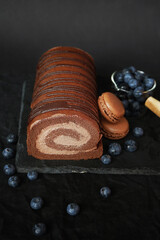 Chocolate roll with chocolate cream next to chocolate macaroons on a black tray next to blueberries on a dark background