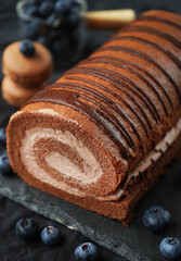 Chocolate roll with chocolate cream next to chocolate macaroons on a black tray next to blueberries on a dark background