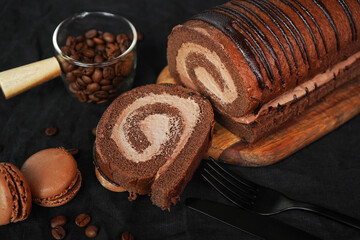 Chocolate roll with chocolate cream next to chocolate macaroons on a black tray next to coffee beans on a dark background