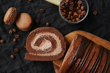 Chocolate roll with chocolate cream next to chocolate macaroons on a black tray next to coffee beans on a dark background
