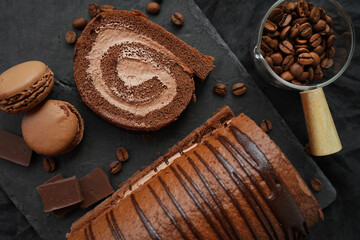 Chocolate roll with chocolate cream next to chocolate macaroons on a black tray next to coffee beans on a dark background