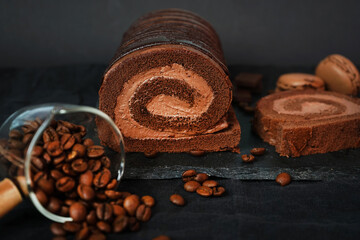 Chocolate roll with chocolate cream next to chocolate macaroons on a black tray next to coffee beans on a dark background