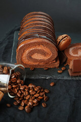 Chocolate roll with chocolate cream next to chocolate macaroons on a black tray next to coffee beans on a dark background