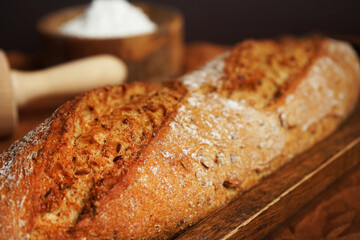 Fresh bread on a wooden tray and a brown napkin next to a bowl of flour and a wooden rolling pin on a brown background