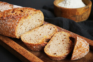Fresh sliced ​​bread on a wooden tray next to a bowl of flour and a wooden rolling pin on a dark background