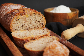 Fresh sliced ​​bread on a wooden tray next to a bowl of flour and a wooden rolling pin on a dark background