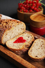 Fresh sliced ​​bread with a heart of berry jam on a wooden tray next to a bowl of red currants and jam on a dark background