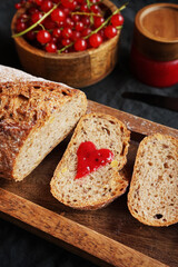 Fresh sliced ​​bread with a heart of berry jam on a wooden tray next to a bowl of red currants and jam on a dark background