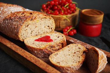 Fresh sliced ​​bread with a heart of berry jam on a wooden tray next to a bowl of red currants and jam on a dark background