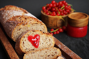 Fresh sliced ​​bread with a heart of berry jam on a wooden tray next to a bowl of red currants and jam on a dark background