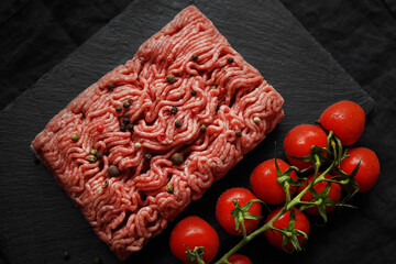 Minced meat with black and red peppers on a black slate tray next to a branch of tomatoes on a dark...