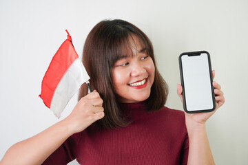 Naklejka premium Smiling young asian woman is showing vertical blank smartphone screen for advertising and holding small Indonesian flag in her hand, isolated over white background. Indonesian Independence Day.