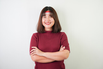 Excited young asian woman wearing Indonesian flag headband, smiling expression with arms crossed on chest gesture, isolated over white background. Concept for Indonesian Independence Day.