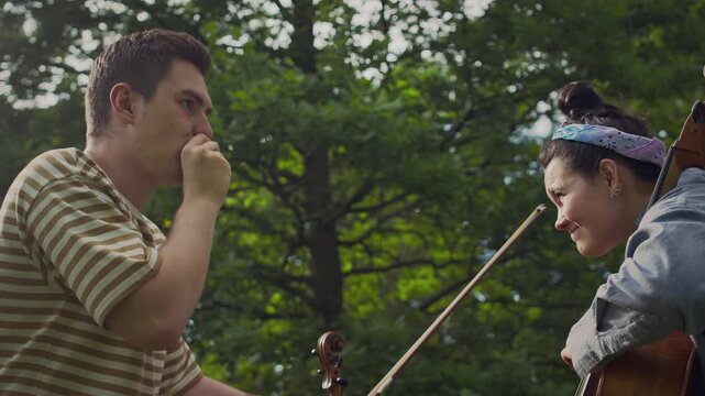 Young cheerful girl playing cello and her creative friend beatboxing while having fun during outdoor performance in park
