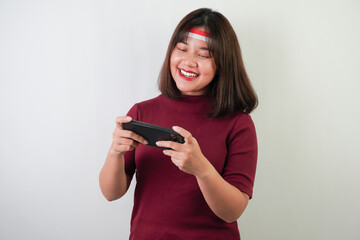 Excited young asian woman wearing Indonesian Flag headband is playing mobile games using smartphone, isolated over white background. Indonesian Independence Day.