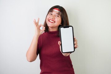 Naklejka premium Smiling young asian woman wearing Indonesian Flag headband is showing vertical blank smartphone screen for advertising with okay gesture, isolated over white background. Indonesian Independence Day.