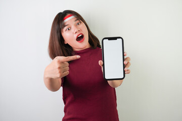 Fototapeta premium Surprised amazed young asian woman wearing Indonesian Flag headband is pointing vertical blank smartphone screen for advertising, isolated over white background. Indonesian Independence Day.