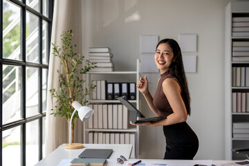 A woman is standing in front of a desk with a laptop and a pen