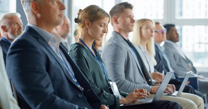 Young Woman Sitting in Crowded Audience at a Business Conference. Female Delegate Smiling and Using Laptop Computer. Manager Watching Motivational Presentation About Investing In Innovative Startups.