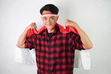 Energetic young asian man is holding and drapes Indonesian flag over his shoulders and back, excited expression, isolated over white background. Concept for Indonesian Independence Day.