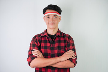 Excited young asian man wearing Indonesian flag headband, smiling expression with arms crossed on chest gesture, isolated over white background. Concept for Indonesian Independence Day.