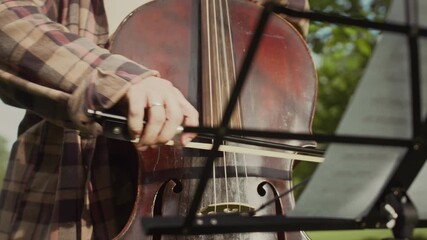 Close-up view of musician playing cello with bow in outdoor park in front of sheet music stand