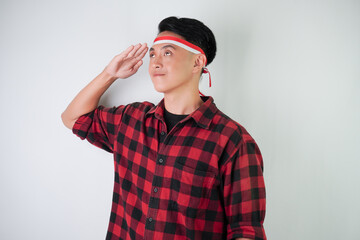 Excited young asian man wearing Indonesian flag headband, smiling expression with saluting gesture, isolated over white background. Concept for Indonesian Independence Day.