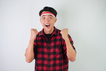 Excited young asian man wearing Indonesian flag headband, smiling expression, hands raised with clenched fist gesture, isolated over white background. Concept for Indonesian Independence Day.