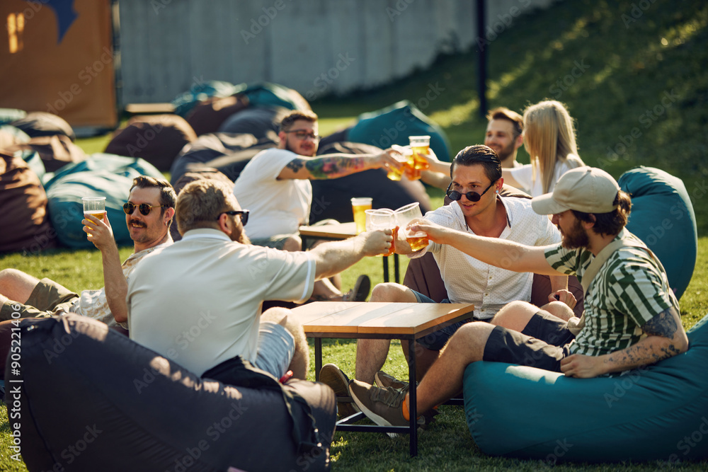 Wall mural Relaxing moment, Happy young people, friends gathering outdoors for picnic, sitting on bean bag chairs, drinking beer and enjoying friendly company. Concept of summer, leisure, friendship, relaxation