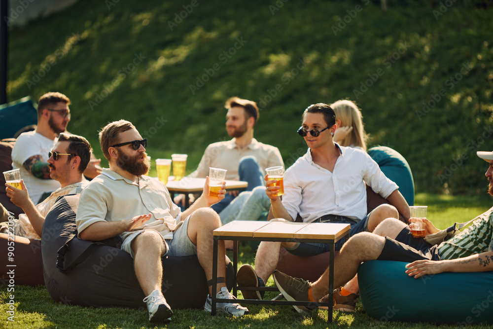 Wall mural Involving conversation. Young people, men sitting on beanbag chair, enjoying outdoor picnic, drinking beer and cheerfully talking. Concept of summer, leisure, friendship, meeting, fun, relaxation