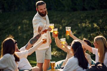 Group of happy smiling young people, friends gathering on outdoor picnic, drinking refreshing drink, having good company. Concept of summer, leisure, friendship, meeting, fun, relaxation