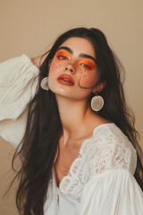 A young woman with long dark hair and vibrant orange eye makeup, wearing a white blouse and large earrings, posing with a serene expression against a beige background