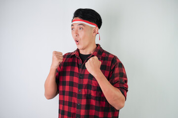 Excited young asian man wearing Indonesian flag headband, smiling expression, hands raised with clenched fist gesture, isolated over white background. Concept for Indonesian Independence Day.