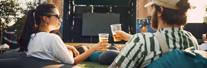 Man and woman, friends gathering outdoor to watch movie on warm summer day, clinking beer glasses and enjoying relaxing vibes. Concept of summer, leisure, friendship, meeting, fun, relaxation