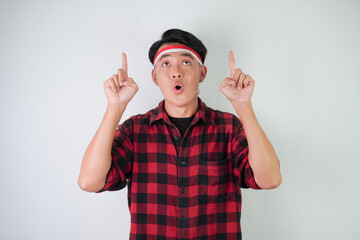 Surprised amazed young asian man wearing Indonesian flag headband and pointing fingers on copy space upside, isolated over white background. Concept for Indonesian Independence Day.