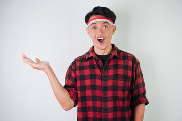 Surprised young asian man wearing Indonesian flag headband, amazed expression with hands open gesture carrying something, isolated over white background. Concept for Indonesian Independence Day.
