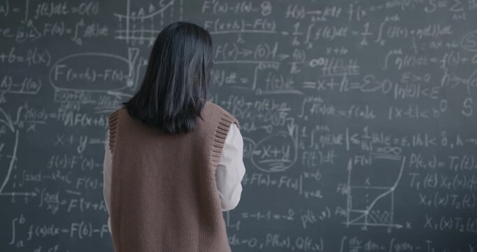 Slow motion back view of Asian professor walking to chalkboard thinking about mathematics theory then underlining formula. Education and research concept.