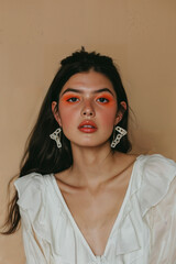 A young woman with long dark hair and vibrant orange eye makeup, wearing a white blouse and large earrings, posing with a serene expression against a beige background